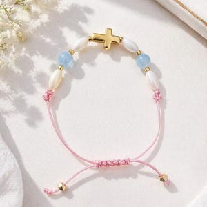 Puede incluir: Una pulsera con un dije de cruz en tono dorado central, flanqueado por cuentas ovaladas blancas y cuentas redondas azul claro. La pieza está ensartada en un cordón rosa con nudos deslizantes ajustables y termina con pequeñas cuentas doradas.