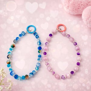 May include: Two beaded necklaces. One features blue and multicoloured beads with a blue clasp. The other has pink and purple beads with a pink clasp. The necklaces are on a pink background.