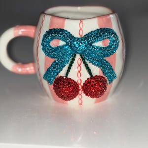 Könnte beinhalten: Eine Keramiktasse mit weißem Henkel und einem rosa-weiß gestreiften Muster. Die Tasse ist mit einer blauen, mit Juwelen besetzten Schleife und zwei roten, mit Juwelen besetzten Kirschen verziert. Die Tasse ist etwa 7,6 cm hoch.