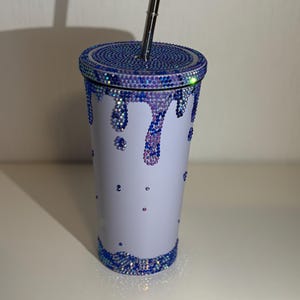 Könnte beinhalten: Ein weißer Becher mit Deckel und silbernem Strohhalm. Der Deckel und der Boden sind mit blauen Strasssteinen besetzt. Blaue und violette Strasssteine ​​tropfen an der Seite des Bechers herunter, mit zusätzlichen Strasssteinen, die auf dem Körper verstreut sind.