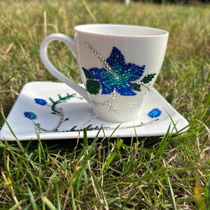 Könnte beinhalten: Weißes Tee-Set mit Blumendesign. Die Tasse hat eine große blau-grüne Blume mit Strasssteinen. Die passende Untertasse hat ein ähnliches Design mit kleineren Blumen und silbernen Akzenten. Das Set steht auf grünem Gras.