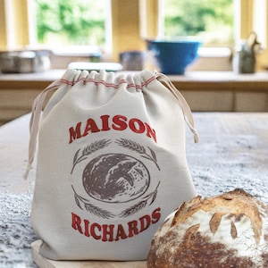 May include: A beige drawstring bread bag with the words "MAISON RICHARDS" in red, and a wheat and bread graphic. A loaf of bread sits next to the bag on a wooden surface.