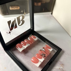 Könnte beinhalten: Ein Set künstlicher Nägel in einer schwarzen Präsentationsbox. Die Nägel haben ein French-Maniküre-Design mit pfirsichfarbener Basis und weißen Spitzen. Einige Nägel sind mit silbernen und goldenen Details verziert. Auf der Box stehen die Buchstaben "VB".