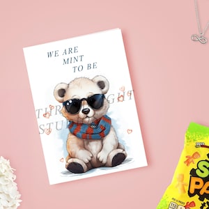 May include: A greeting card featuring a cartoon bear wearing sunglasses and a scarf. The card reads "WE ARE MINT TO BE". The bear is sitting on a light blue background with small hearts. A white flower arrangement and a bag of candy are in the background.