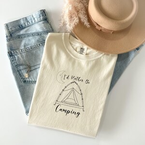 Peut inclure: T-shirt crème avec l'inscription "I'd Rather Be Camping" et un dessin de tente. Le t-shirt est associé à un jean délavé, un chapeau beige et un collier. Une brindille de fleurs séchées ajoute une touche décorative.
