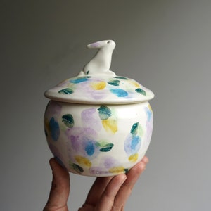 May include: White ceramic jar with a lid featuring a small white rabbit figurine. The jar is decorated with a colorful floral pattern in shades of purple, blue, yellow, and green.