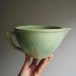 May include: A light green ceramic mixing bowl with a handle and a pouring spout. The bowl has a ribbed texture and is held by a hand against a neutral background. The bowl is a vintage kitchenware item.