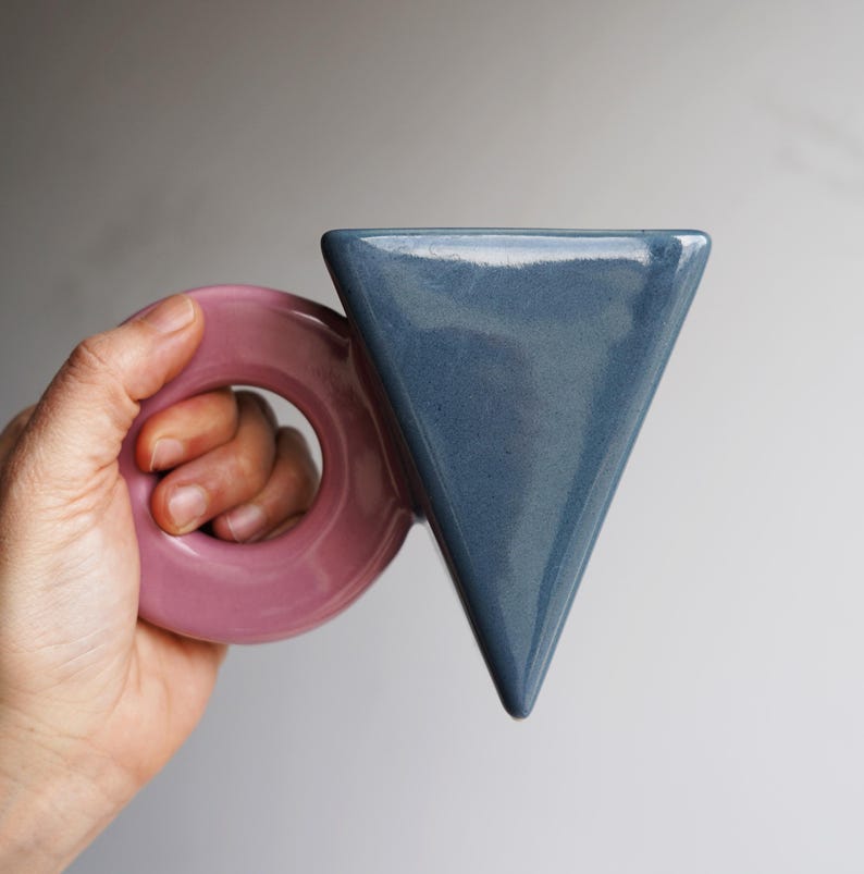 May include: A distinctive ceramic mug with a geometric design. The mug has a triangular body in a dusty blue colour and a circular handle in a soft pink hue. The mug is held in a hand against a neutral background.