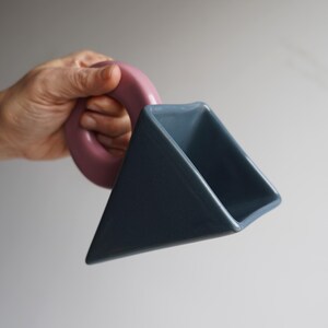 May include: A distinctive ceramic mug with a geometric design. The mug has a triangular body in a muted blue-grey colour and a circular handle in a dusty rose hue. The mug's design is modern and artistic, suitable for beverages.