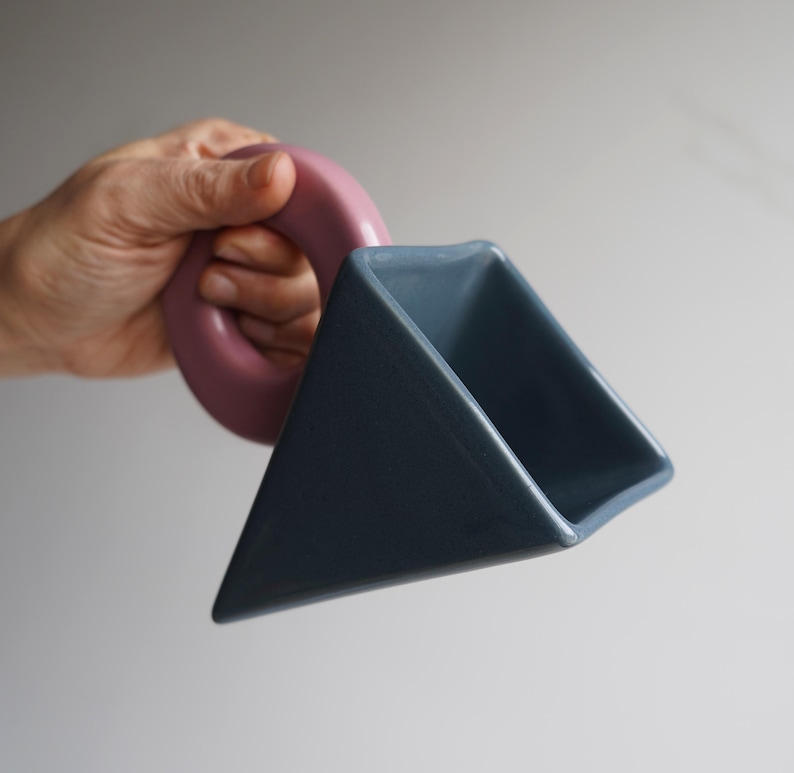 May include: A distinctive ceramic mug with a geometric design. The mug has a triangular body in a muted blue-grey colour and a circular handle in a dusty rose hue. The mug's design is modern and artistic, suitable for beverages.