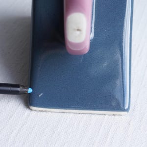 May include: A close-up of a blue ceramic soap dish with a pink handle and a white accent. A blue pencil is positioned near the corner of the dish, suggesting scale and use. The dish is on a white, textured surface.