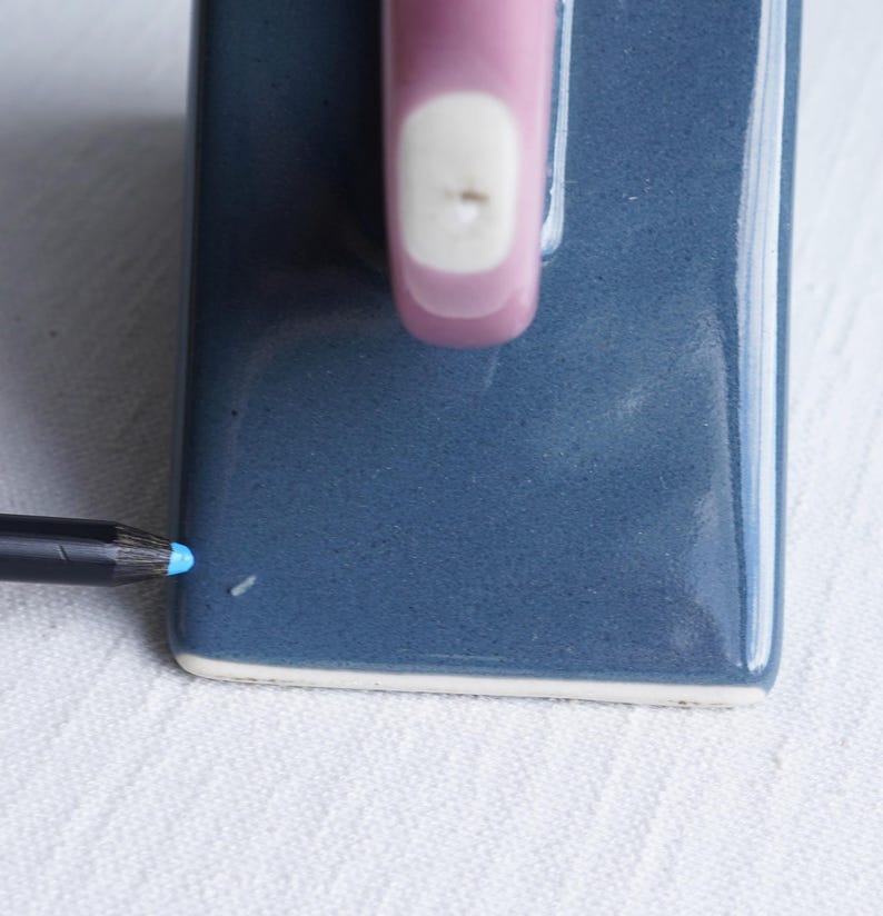 May include: A close-up of a blue ceramic soap dish with a pink handle and a white accent. A blue pencil is positioned near the corner of the dish, suggesting scale and use. The dish is on a white, textured surface.