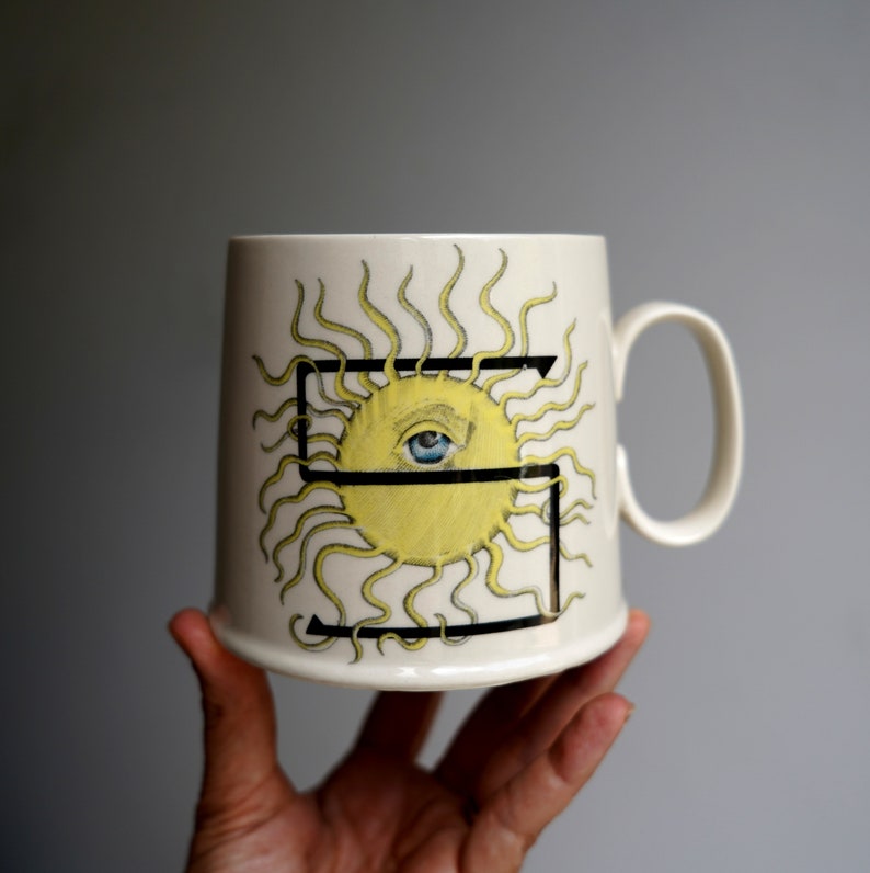 K&ouml;nnte beinhalten: Cremefarbene Keramiktasse mit gelbem Sonnendesign, blauem Auge und schwarzem Rahmen. Die Sonne hat strahlende gelbe Linien. Die Tasse hat einen gebogenen Henkel und eine zylindrische Form.