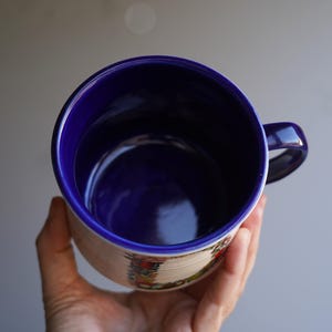 K&ouml;nnte beinhalten: Eine Keramiktasse mit blauem Innenraum und Henkel, gehalten in einer Hand. Die Au&szlig;enseite ist cremefarben mit einem dekorativen Band aus bunten Blumenmustern. Der Rand und der Henkel der Tasse sind tiefblau.
