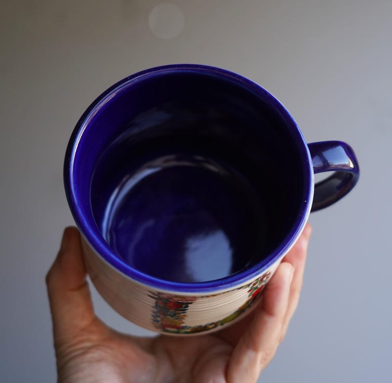 K&ouml;nnte beinhalten: Eine Keramiktasse mit blauem Innenraum und Henkel, gehalten in einer Hand. Die Au&szlig;enseite ist cremefarben mit einem dekorativen Band aus bunten Blumenmustern. Der Rand und der Henkel der Tasse sind tiefblau.