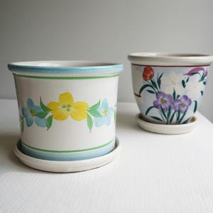 May include: Two ceramic plant pots with matching saucers. The front pot is white with a horizontal band of painted yellow and blue flowers. The back pot features a painted floral arrangement with purple, white, and red blossoms on a white base.
