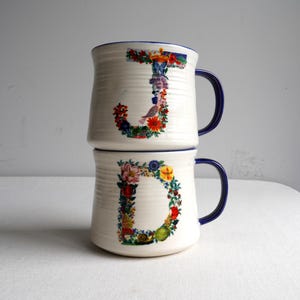 K&ouml;nnte beinhalten: Zwei gestapelte wei&szlig;e Keramikbecher mit blauen Griffen und R&auml;ndern. Der Buchstabe "J" ist mit bunten Blumen und einem Vogel auf dem oberen Becher verziert, und der Buchstabe "D" ist &auml;hnlich auf dem unteren Becher verziert. Die Becher haben eine gerippte Textur.