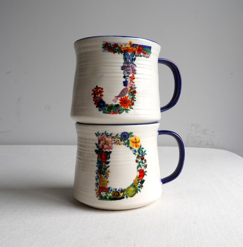 K&ouml;nnte beinhalten: Zwei gestapelte wei&szlig;e Keramikbecher mit blauen Griffen und R&auml;ndern. Der Buchstabe "J" ist mit bunten Blumen und einem Vogel auf dem oberen Becher verziert, und der Buchstabe "D" ist &auml;hnlich auf dem unteren Becher verziert. Die Becher haben eine gerippte Textur.