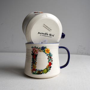 K&ouml;nnte beinhalten: Zwei wei&szlig;e Keramikbecher mit blauen Henkeln und R&auml;ndern. Einer der Becher zeigt ein gro&szlig;es Blumenmuster in Form eines "D". Der andere Becher ist umgedreht und zeigt die Marke "Nathalie Lete for Anthropologie" auf der Unterseite.