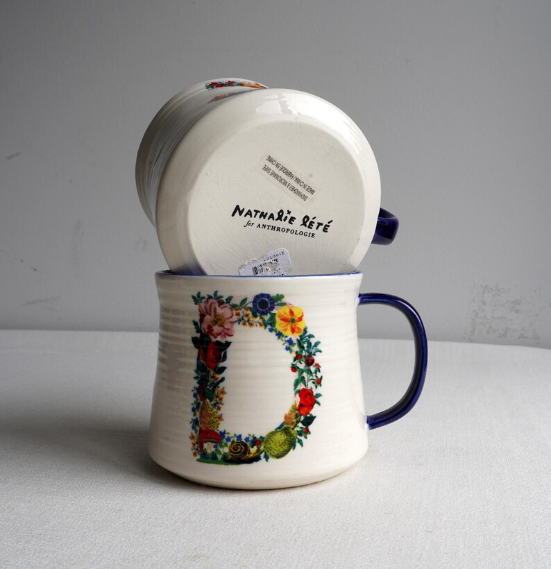 K&ouml;nnte beinhalten: Zwei wei&szlig;e Keramikbecher mit blauen Henkeln und R&auml;ndern. Einer der Becher zeigt ein gro&szlig;es Blumenmuster in Form eines "D". Der andere Becher ist umgedreht und zeigt die Marke "Nathalie Lete for Anthropologie" auf der Unterseite.