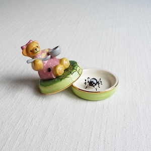 May include: A whimsical ceramic trinket box featuring a yellow teddy bear with a pink dress and a spoon, resting on a green base with a spiderweb design. The open box reveals a small spider figurine inside. The box has a gold trim.