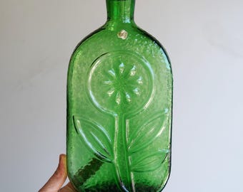 May include: A green glass bottle with a textured surface and a flower design. The bottle has a rectangular shape with rounded edges and a narrow neck. The flower design is embossed on the front of the bottle.