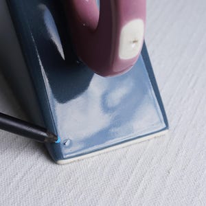 May include: A ceramic mug with a rectangular base and a rounded handle. The mug is a deep blue colour with a mauve handle and a white accent. A black pencil with a blue tip is touching the mug.