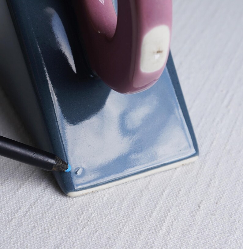 May include: A ceramic mug with a rectangular base and a rounded handle. The mug is a deep blue colour with a mauve handle and a white accent. A black pencil with a blue tip is touching the mug.