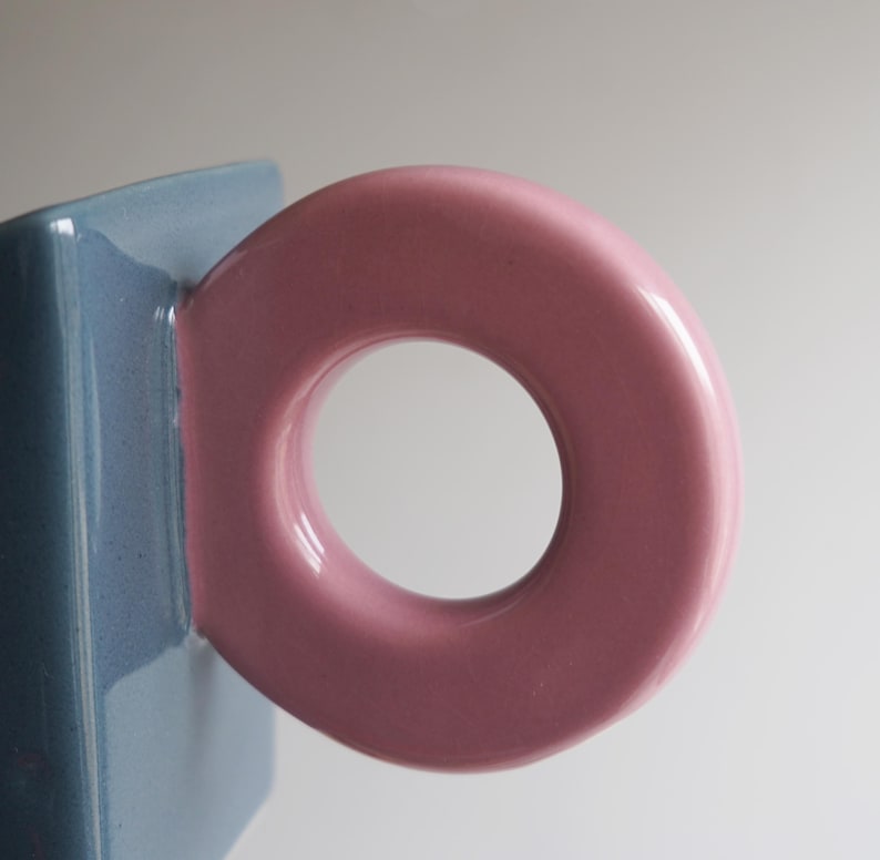 May include: A close-up shot of a ceramic mug with a distinctive design. The mug has a light blue body and a large, circular, pink handle. The handle's shape is a key design element, contrasting with the mug's angular form.