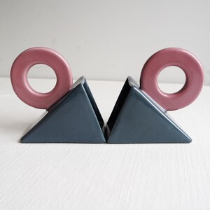 May include: A pair of ceramic bookends. Each bookend features a dark blue triangular base with a pink, donut-shaped accent. These bookends are designed to hold books upright on a shelf or desk.