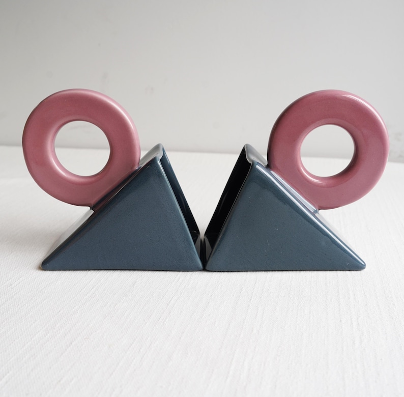 May include: A pair of ceramic bookends. Each bookend features a dark blue triangular base with a pink, donut-shaped accent. These bookends are designed to hold books upright on a shelf or desk.