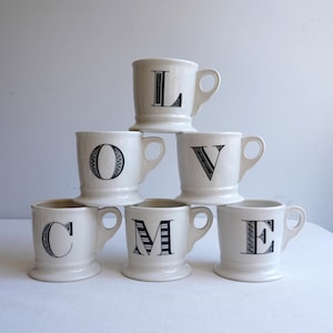 Pu&ograve; includere: Sei tazze in ceramica bianca con disegni di lettere nere che compongono la parola "LOVE" impilate sopra la parola "COME".