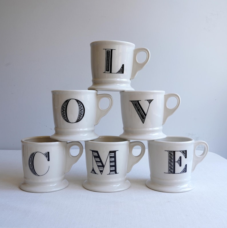 Pu&ograve; includere: Sei tazze in ceramica bianca con disegni di lettere nere che compongono la parola "LOVE" impilate sopra la parola "COME".