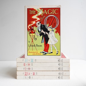 May include: A stack of vintage books, including "The Magic of Oz" by L. Frank Baum, illustrated by John R. Neill. The top book features a red and yellow cover illustration. The other books have white covers with the title "Oz" and the Rand McNally logo.
