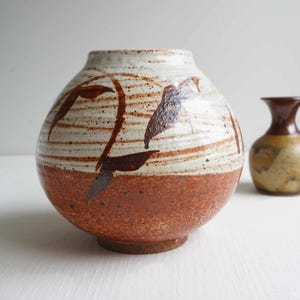 May include: A handcrafted ceramic vase with a rounded shape. The lower half is a reddish-brown color, while the upper half is white with brown leaf-like designs. A smaller vase is visible in the background.
