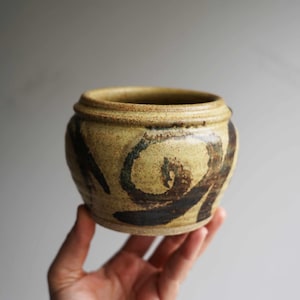 May include: A small, round ceramic pot with a light tan base and a dark brown and teal swirl design. The pot has a slightly textured surface and a rim around the top. The pot is held in a hand.