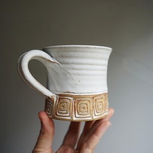 May include: A ceramic mug with a white glazed upper section featuring horizontal ridges and a natural clay base decorated with a repeating pattern of carved brown squares. The mug has a sturdy handle and a flared rim, showcasing a handcrafted design.