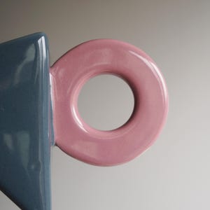 May include: A close-up shot of a ceramic mug with a geometric design. The mug has a blue body and a pink, circular handle. The handle is a key design element, giving a modern look. The mug is likely for drinks.