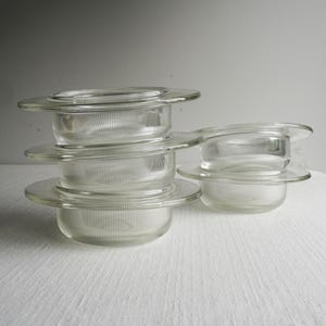 May include: A collection of clear glass candle holders. Each holder has a circular base and a wide, flat rim. The glass has a ribbed texture. The candle holders are stacked and arranged on a white surface.