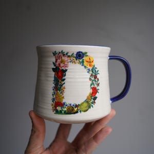 K&ouml;nnte beinhalten: Cremefarbene Keramiktasse mit blauem Henkel und Rand. Der Buchstabe "D" ist mit einem farbenfrohen Blumen- und Fruchtdesign verziert. Die Tasse hat eine strukturierte Oberfl&auml;che und wird von einer Hand gehalten.