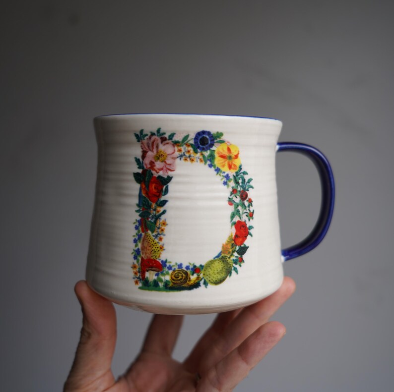 K&ouml;nnte beinhalten: Cremefarbene Keramiktasse mit blauem Henkel und Rand. Der Buchstabe "D" ist mit einem farbenfrohen Blumen- und Fruchtdesign verziert. Die Tasse hat eine strukturierte Oberfl&auml;che und wird von einer Hand gehalten.