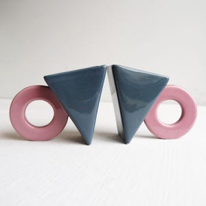 May include: A pair of ceramic bookends. Each bookend has a navy blue triangular shape and a pink circular shape. These decorative bookends are designed to keep books upright on a shelf.
