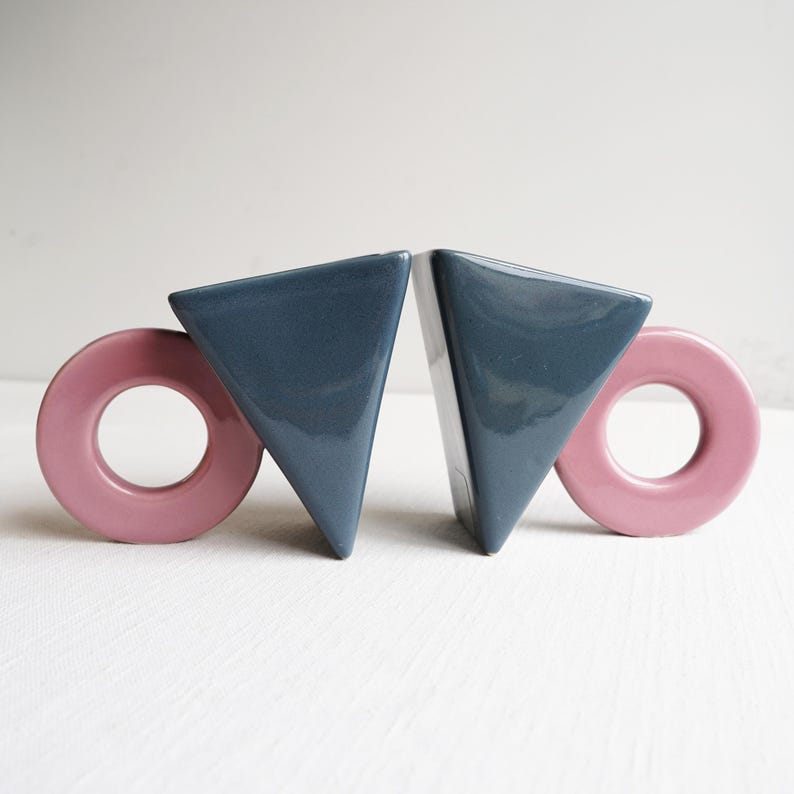 May include: A pair of ceramic bookends. Each bookend has a navy blue triangular shape and a pink circular shape. These decorative bookends are designed to keep books upright on a shelf.
