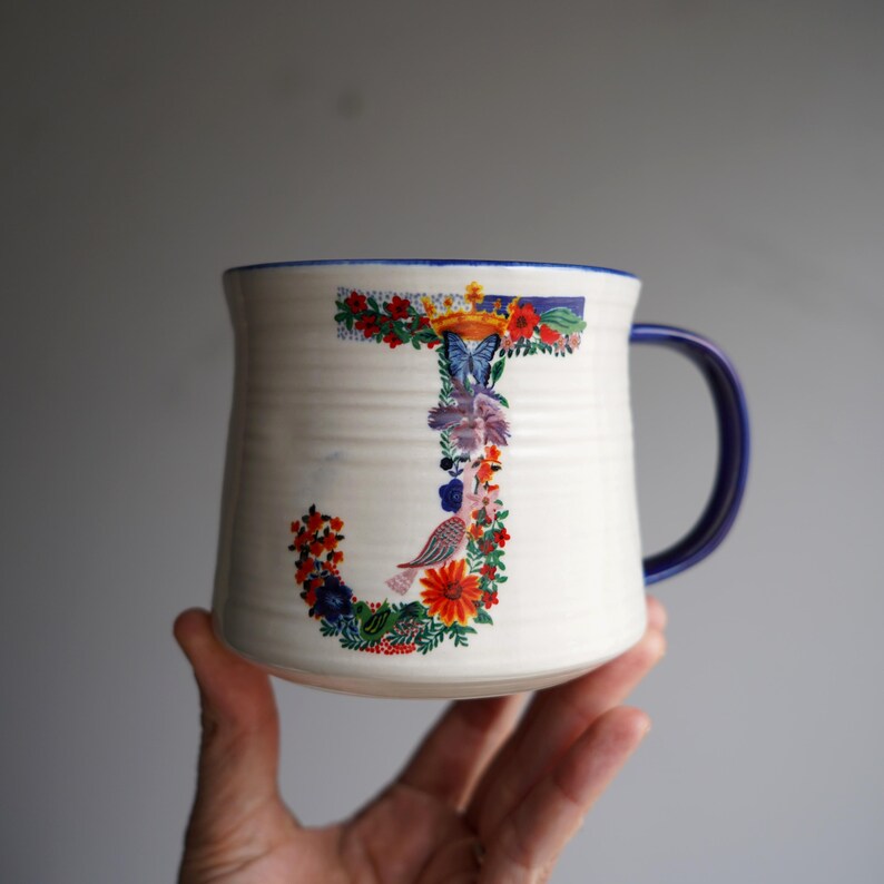 K&ouml;nnte beinhalten: Wei&szlig;er Keramikbecher mit blauem Henkel und Rand. Der Buchstabe &bdquo;J&ldquo; ist mit bunten Blumen, einem Schmetterling und einem Vogel verziert. Der Becher hat eine strukturierte Oberfl&auml;che.
