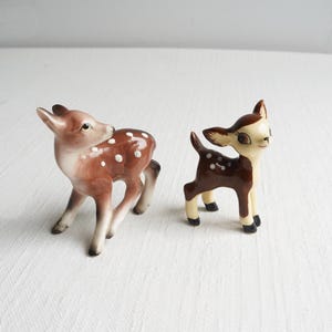 May include: Two ceramic deer figurines. One is brown with white spots and a cream-colored face. The other is a smaller deer with a similar color scheme. Both have black hooves and are standing on a white surface.