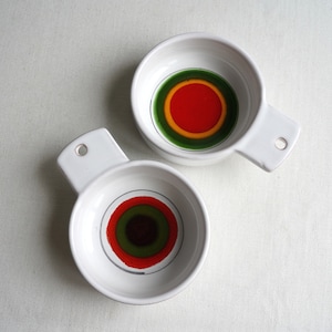 May include: Two white ceramic bowls with handles. The bowls have a concentric circle design in green, yellow, red, and black.