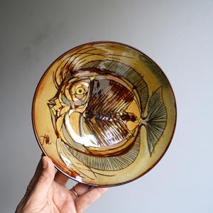 May include: A ceramic plate with a brown and yellow glaze features a hand-painted illustration of a fish with a detailed skeletal structure. The plate is signed "Ha" in the lower left corner.