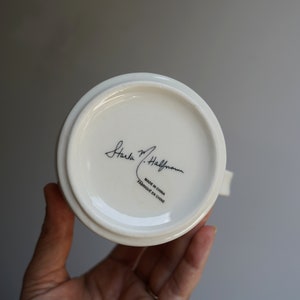 Pu&ograve; includere: Tazza in ceramica bianca con una firma nera che recita "Carla M. Hallstrom" e il testo "Made in China Fabrique en Chine".