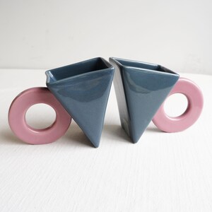 May include: Two ceramic mugs with geometric shapes. The mugs are triangular with a dark blue glaze and have pink, circular handles. The mugs are set against a white background.