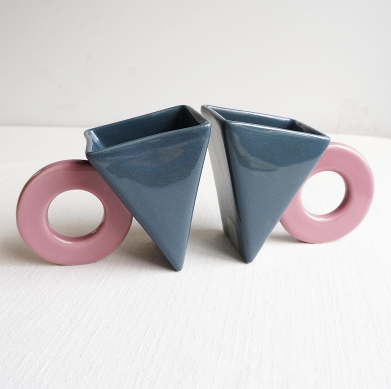 May include: Two ceramic mugs with geometric shapes. The mugs are triangular with a dark blue glaze and have pink, circular handles. The mugs are set against a white background.
